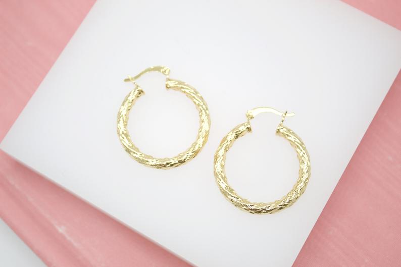 A pair of textured gold hoop earrings rests against a white display surface.