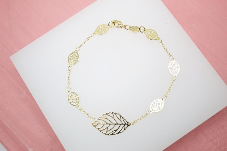 A delicate gold chain bracelet features intricate leaf charms resting on a white display against a soft pink background.