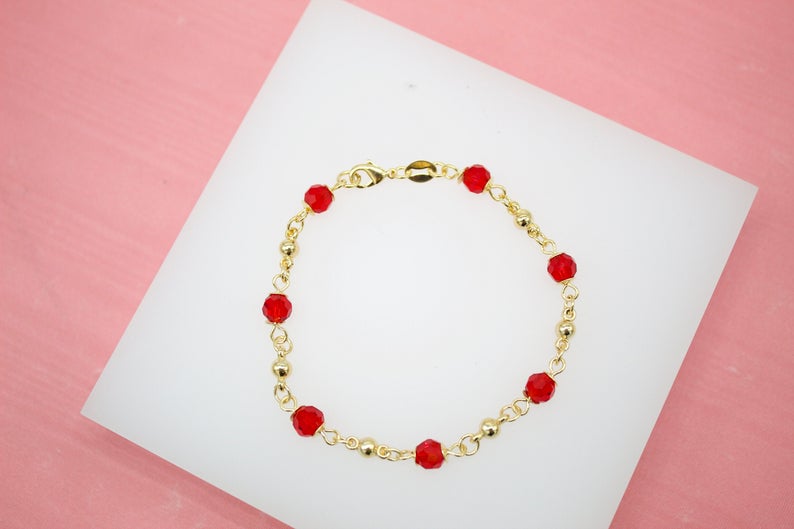 A delicate gold chain bracelet features vibrant red crystal beads resting on a white display surface against a pink background.