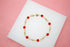 A delicate gold chain bracelet features vibrant red crystal beads resting on a white display surface against a pink background.