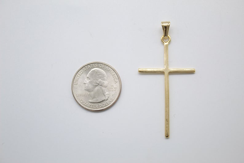 A plain yellow gold cross pendant rests beside a US quarter coin for size comparison on a light surface.