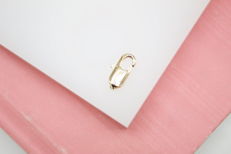 A shiny gold lobster clasp rests on a white display surface bordered by a pink background.