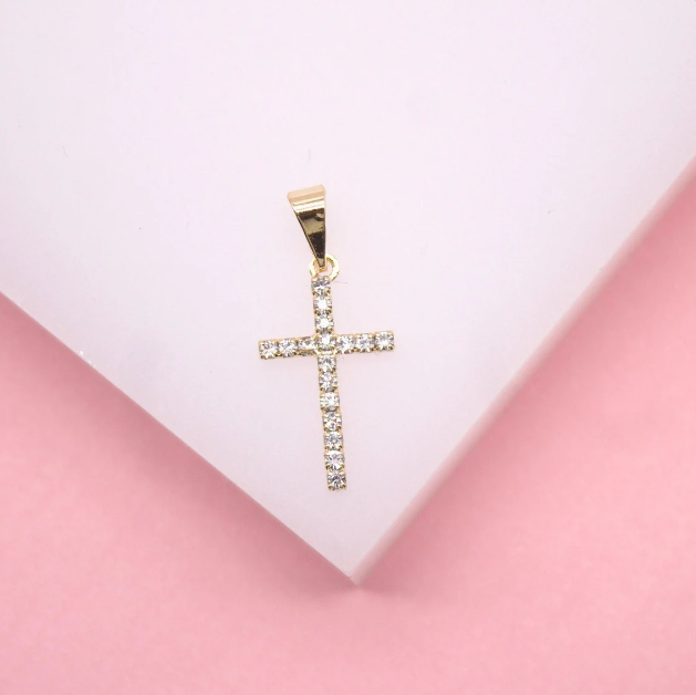 A sparkling gold cross pendant set with small diamonds rests on a light pink surface.