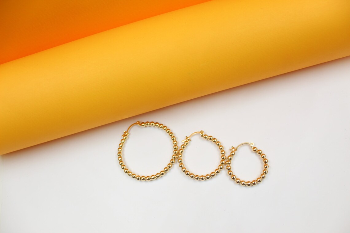 Three graduated sizes of beaded gold hoop earrings rest on a white surface against an orange backdrop.