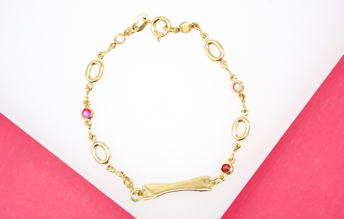 A gold bracelet features alternating oval links and colorful gemstone accents on a pink and white surface.
