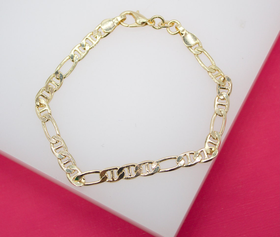 A gold Mariner link bracelet rests on a white display surface against a pink background.