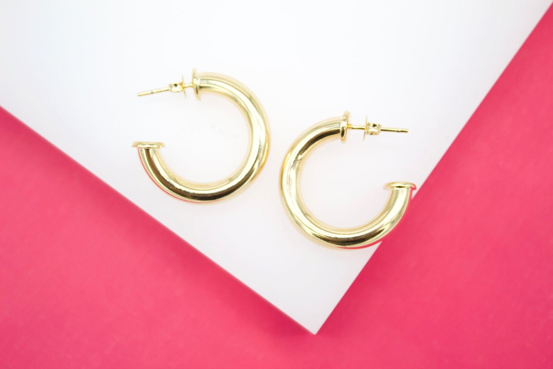 Two thick, polished gold hoop earrings rest on a white surface against a vibrant pink background.