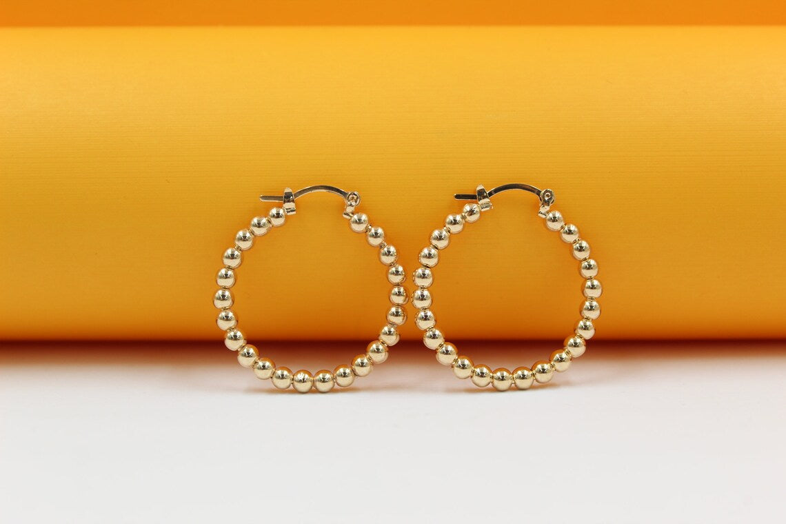 A pair of elegant gold-plated beaded hoop earrings rests against a vibrant yellow and white background.
