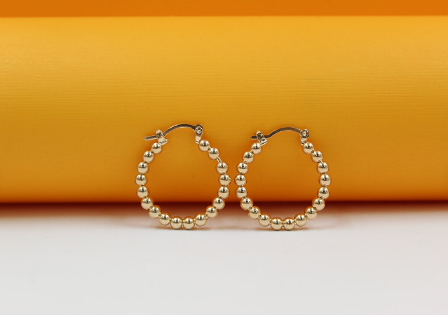 A pair of shiny gold-plated beaded hoop earrings rests against a vibrant orange and white background.