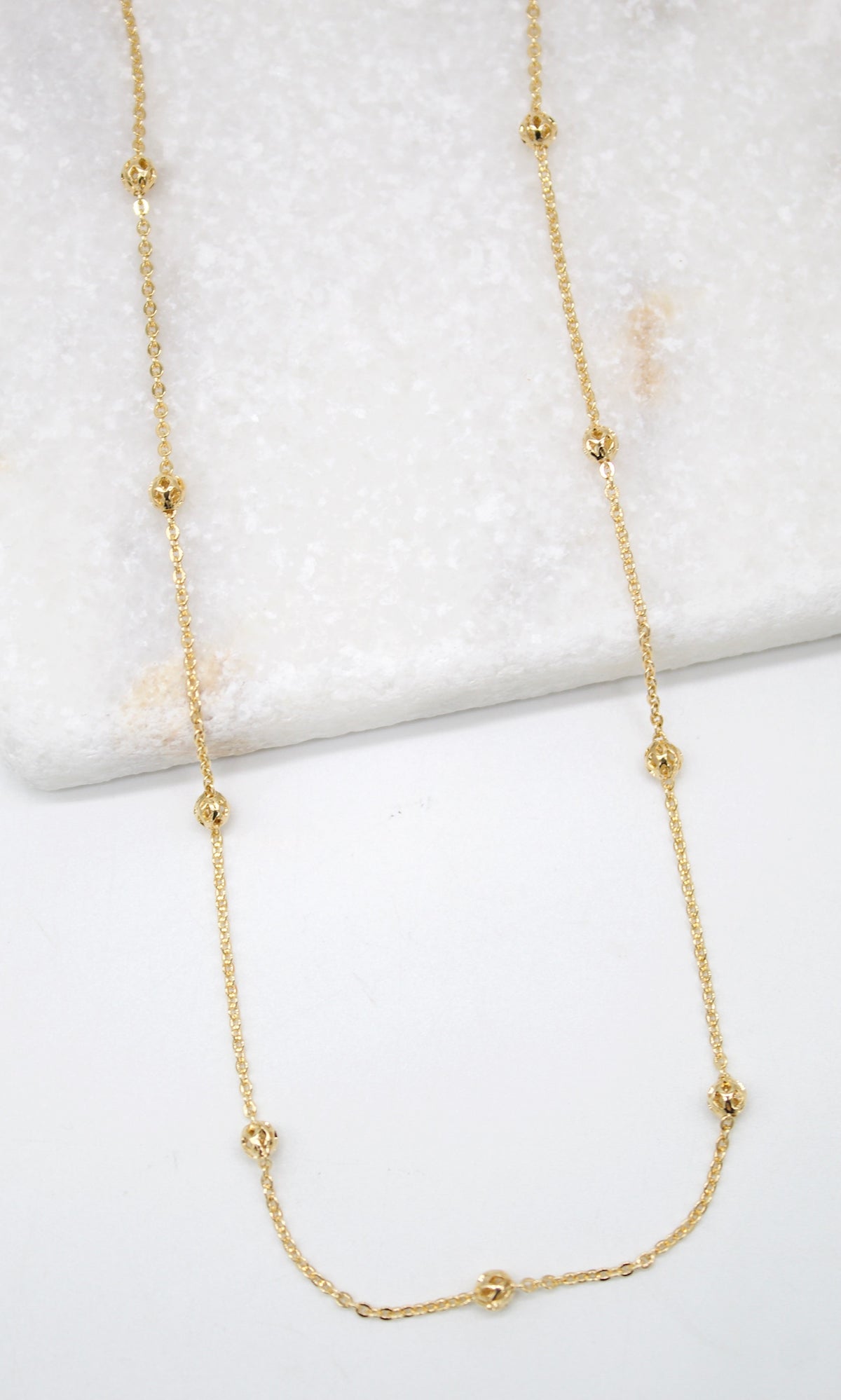 A delicate gold station necklace lies draped against a white marble background.