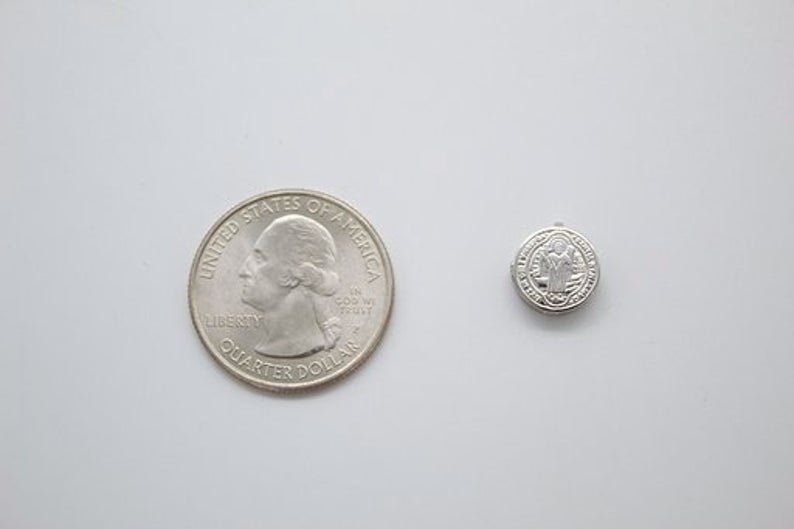 A tiny silver Saint Benedict medal sits next to a US quarter coin for scale on a white surface.