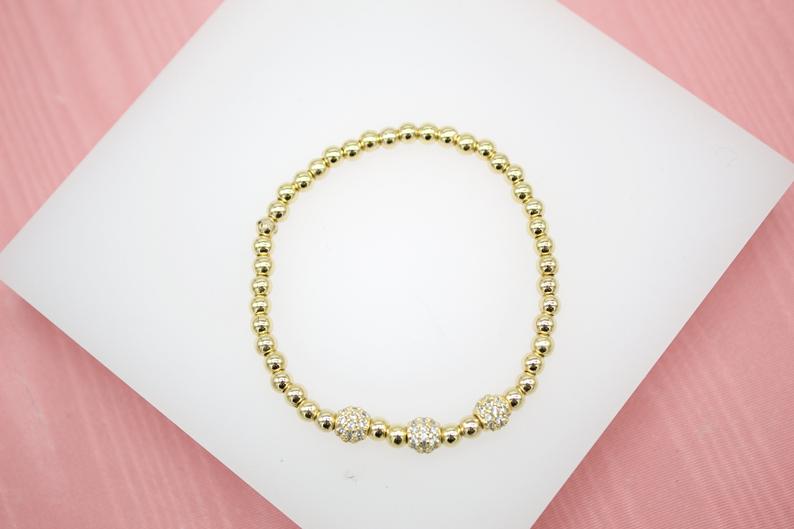 A gold bead stretch bracelet rests on a white display surface, featuring three sparkly pave-set rhinestone disco ball beads.