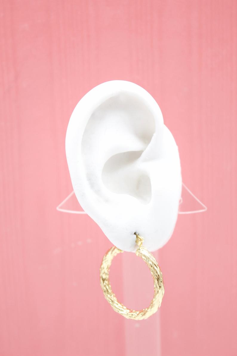 A white mannequin ear displays a textured gold hoop earring against a pink background.