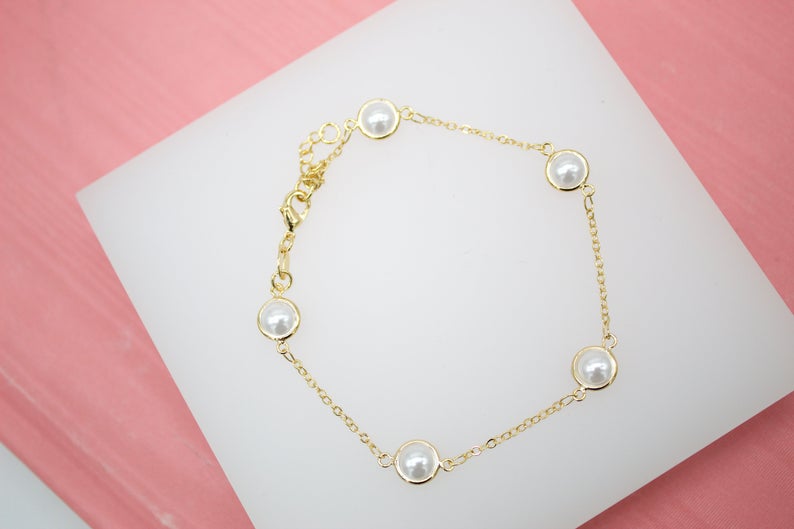 A delicate gold chain bracelet features evenly spaced bezel-set white pearls, resting on a white display against a pink background.