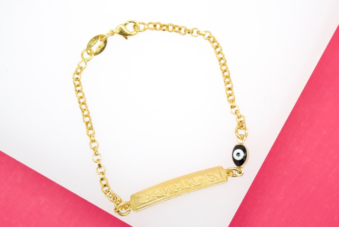 A gold ID bracelet rests on a white and pink surface, featuring an engraved rectangular tag and a protective blue-and-black evil eye charm.