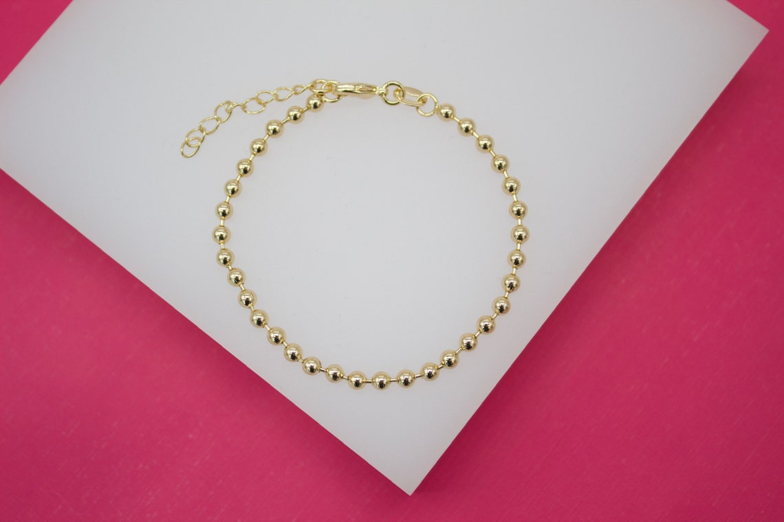 A delicate gold ball chain bracelet rests on a white display against a pink background.