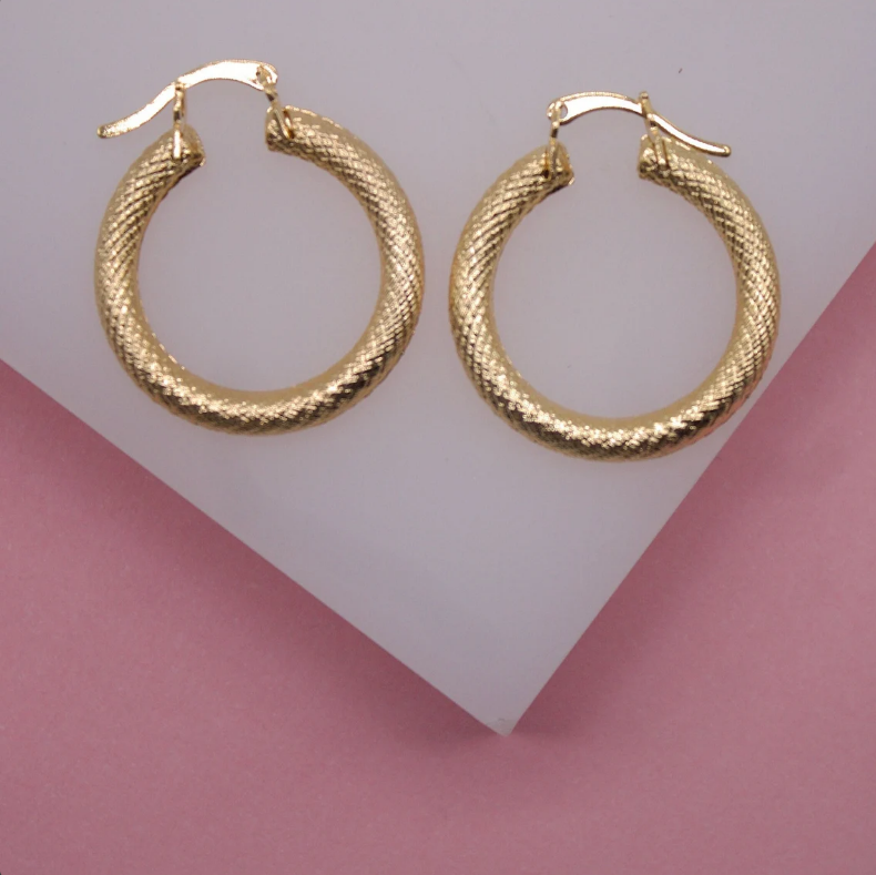 A pair of textured gold hoop earrings rests on a geometric white and pink display.