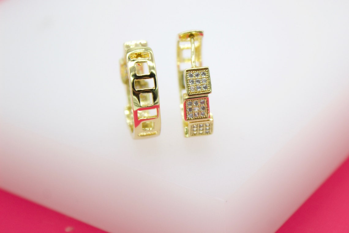 A pair of gleaming gold huggie hoop earrings featuring intricate geometric cutouts and sparkling cubic zirconia pavé accents rests on a clean white surface.