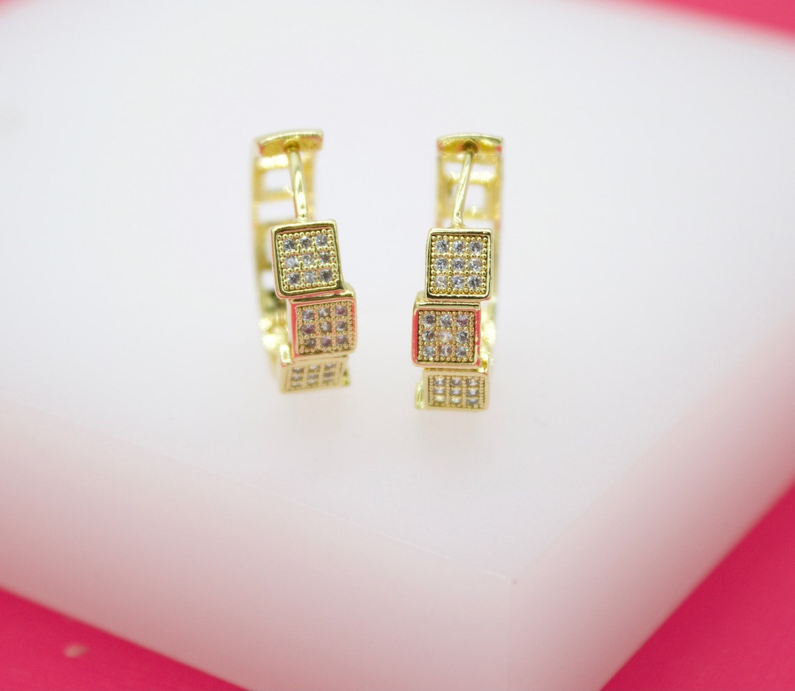 A pair of gold huggie hoop earrings featuring stacked cubic charms encrusted with sparkling micro-pave zirconia stones sits on a white display surface.