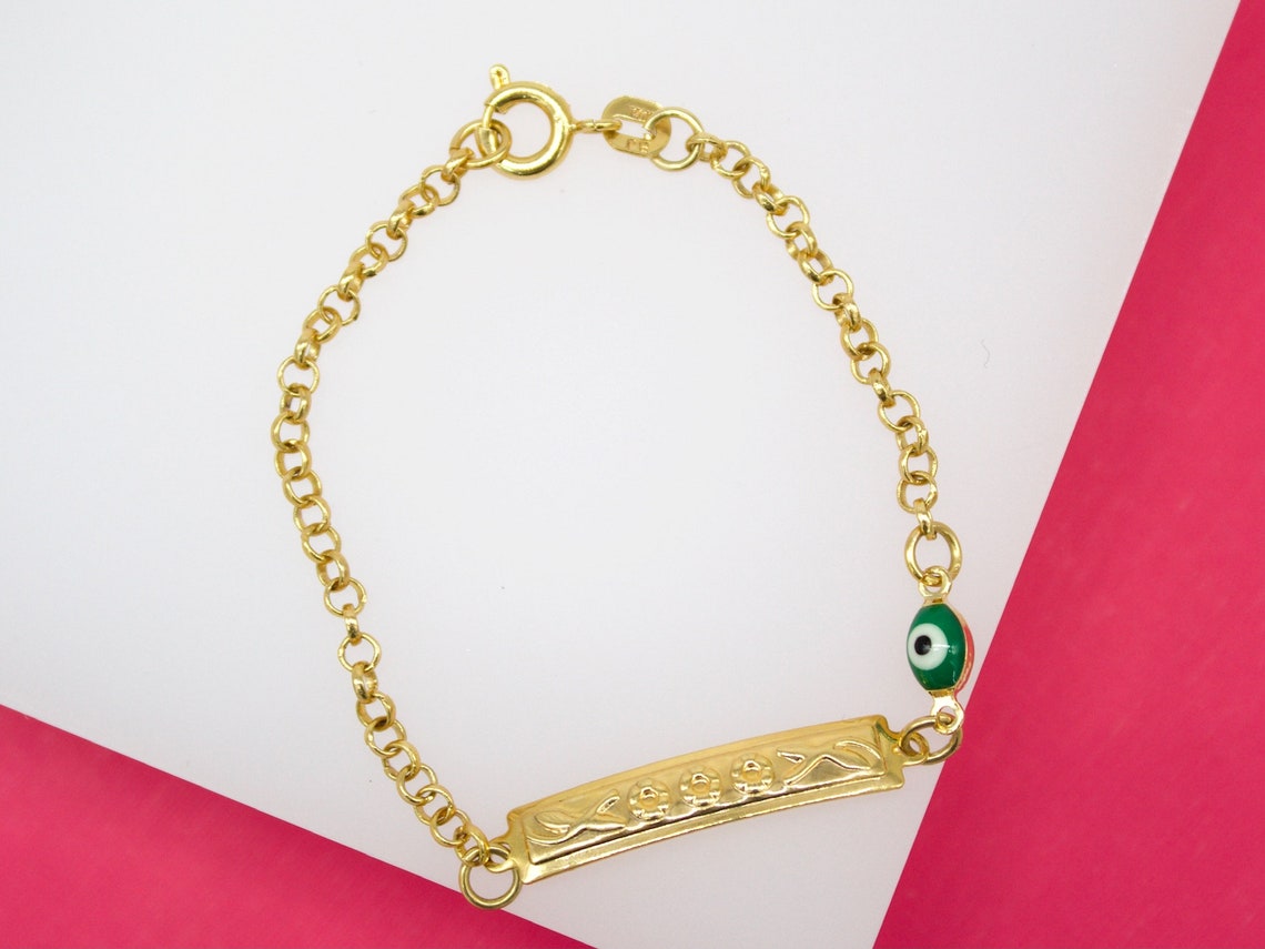 A gold ID bracelet rests on a white surface against a bright pink background, featuring an engraved nameplate and a green evil eye charm.
