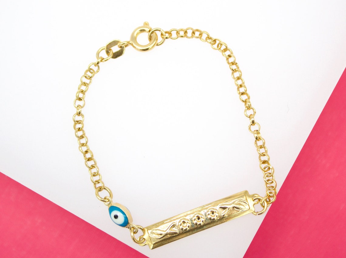 A delicate gold ID bracelet rests on a white and pink background, featuring an engraved rectangular plate and a small blue evil eye charm.