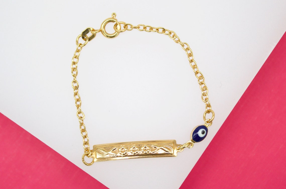 A delicate gold identity bracelet features an engraved bar and a small blue evil eye bead against a white and pink background.
