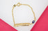 A delicate gold identity bracelet features an engraved bar and a small blue evil eye bead against a white and pink background.
