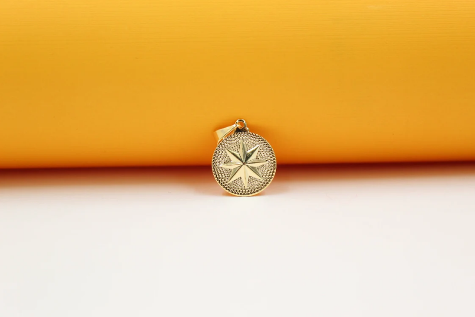 A gold pendant necklace featuring a textured, round compass star charm rests against a vibrant yellow and white background.