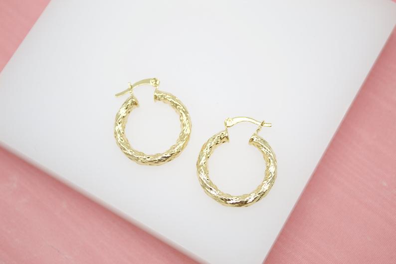 A pair of textured gold hoop earrings rests on a white display surface against a pink background.