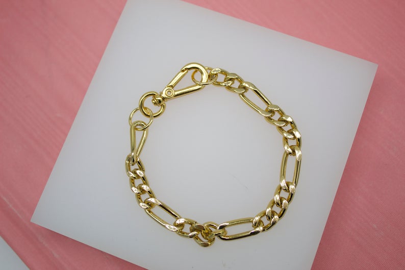 A polished gold Figaro chain bracelet rests on a white display pad against a soft pink background.