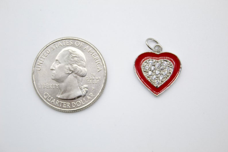 A silver heart-shaped pendant featuring red enamel and sparkling cubic zirconia stones rests beside a US quarter for size comparison on a plain white surface.