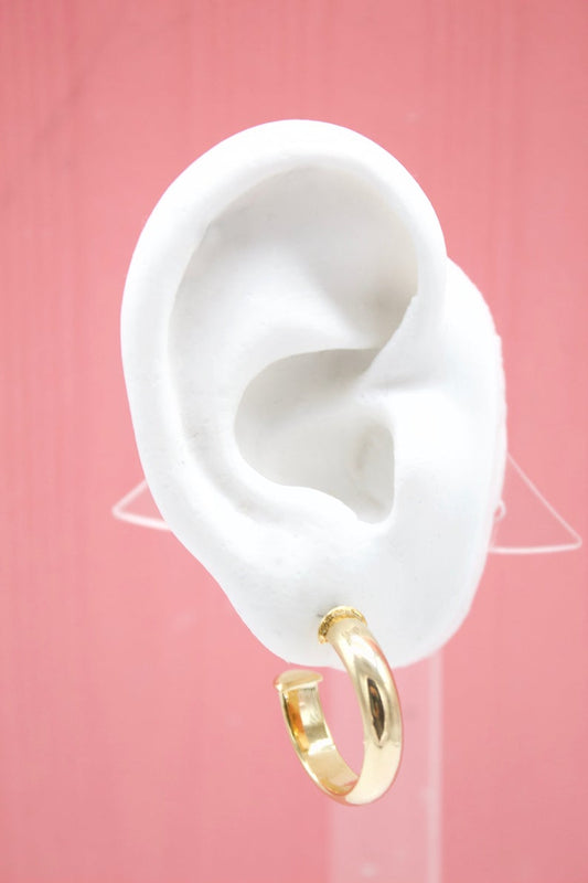 A white mannequin ear displays a polished gold hoop earring against a pink background.