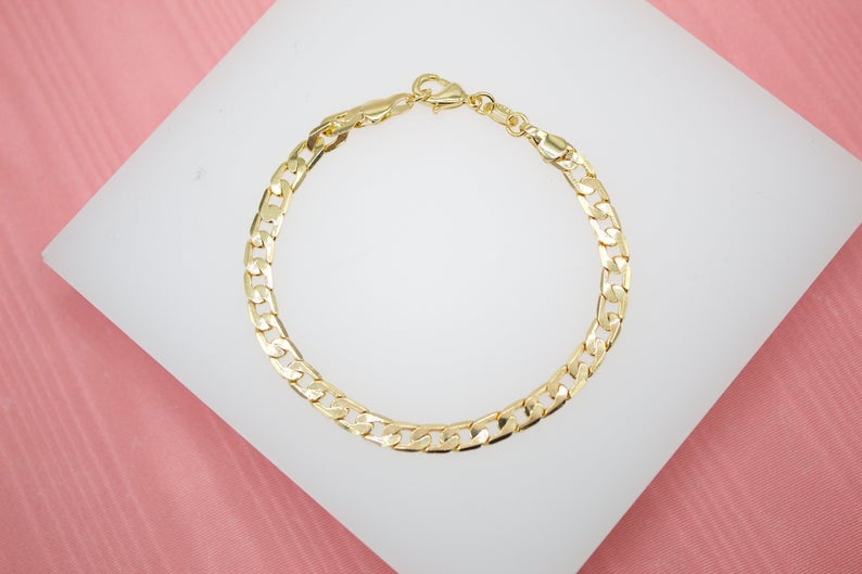 A shiny gold curb chain bracelet rests neatly on a white display stand against a pink background.