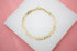 A shiny gold curb chain bracelet rests neatly on a white display stand against a pink background.