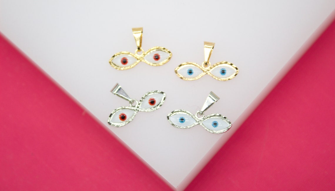 Four double-eyed evil eye charms rest on a white surface, featuring gold and silver finishes with white enamel and colorful red or blue centers.