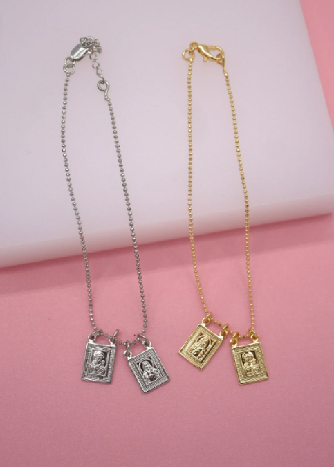 Two delicate scapular bracelets, one silver and one gold ball-chain, lie side by side on a pink and white surface, each featuring two small rectangular religious pendants.