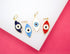 Four colorful enamel evil eye charms in dark blue, light blue, white, and red rest on a white surface against a bright pink background.