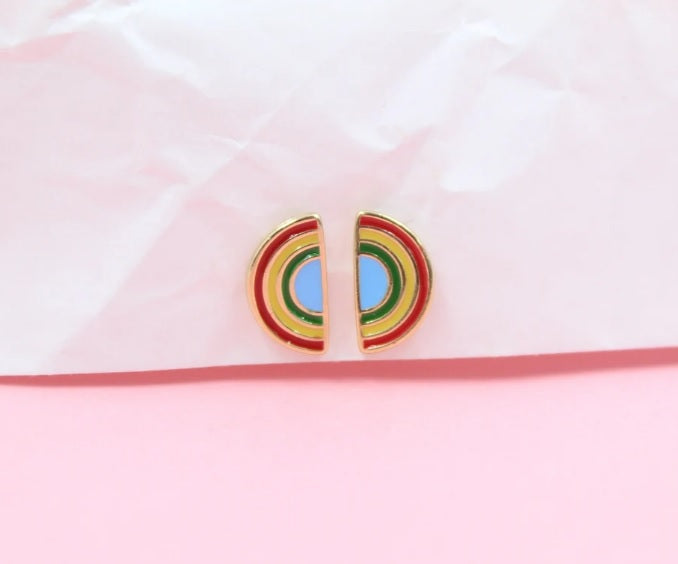 A pair of gold enamel half-circle rainbow stud earrings rests against a crinkled white and pink background.