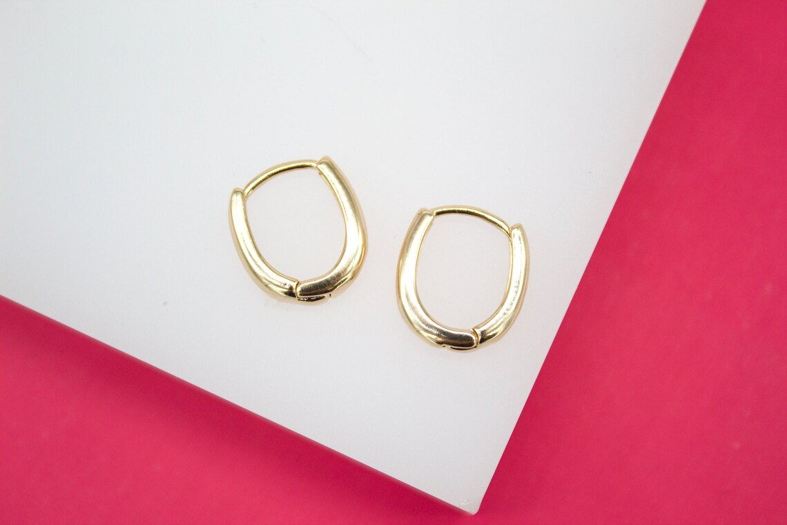 A pair of minimalist gold huggie hoop earrings rests on a white and bright pink display surface.