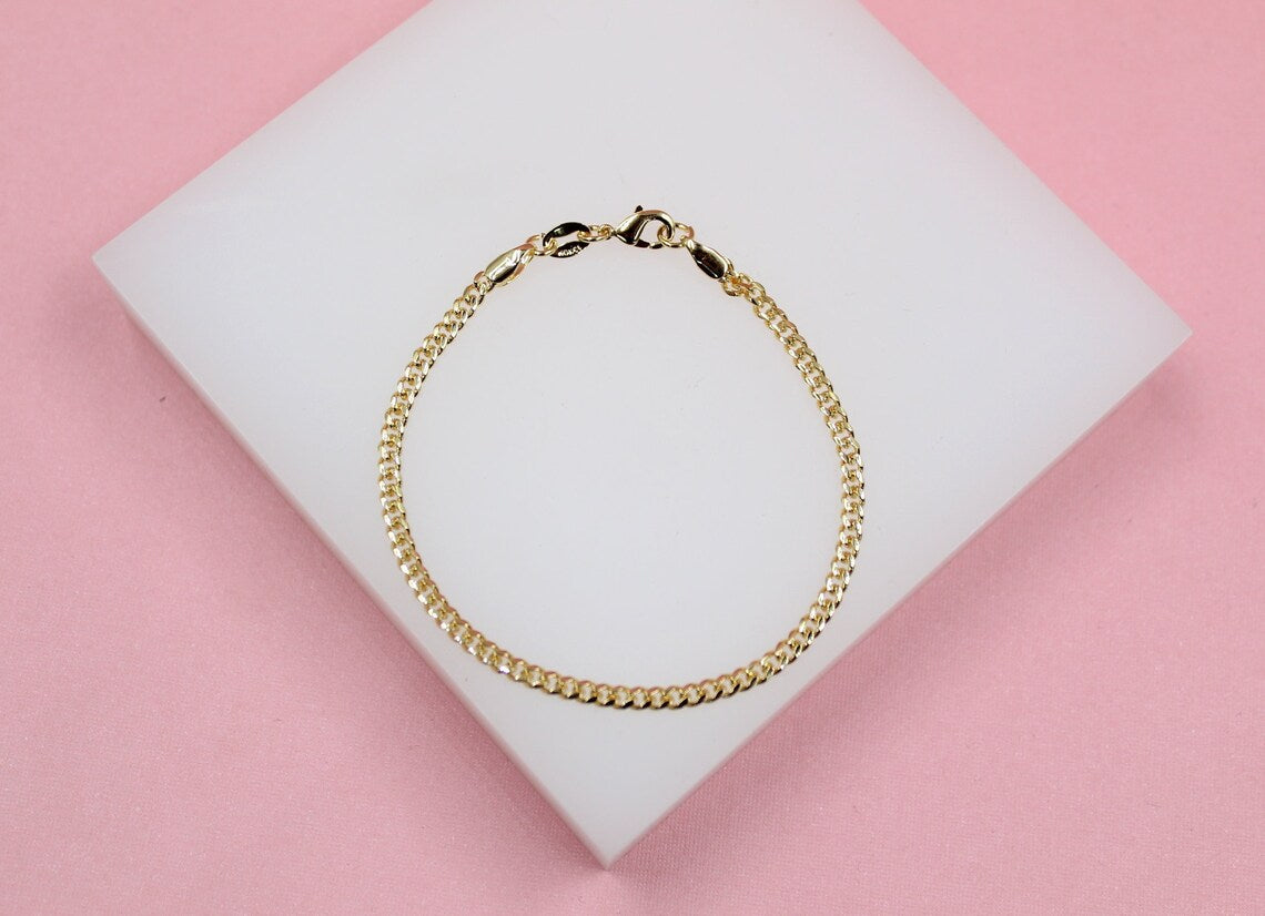 A delicate gold curb chain bracelet rests on a white display block against a soft pink background.