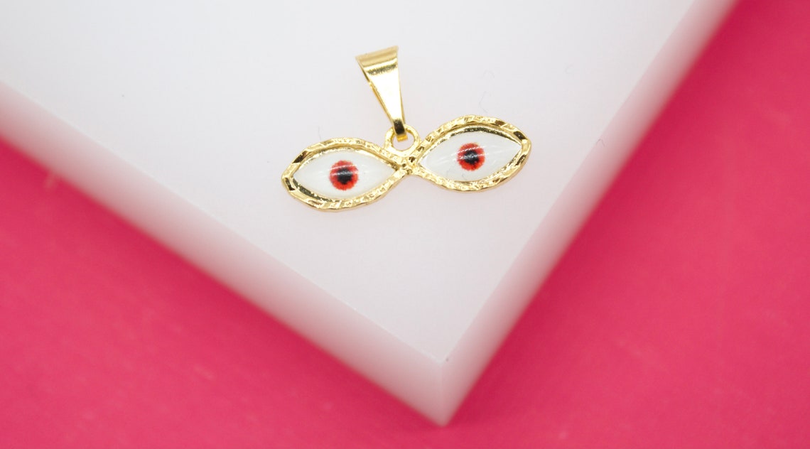 A gold evil eye pendant rests on a white display surface against a vibrant pink background.