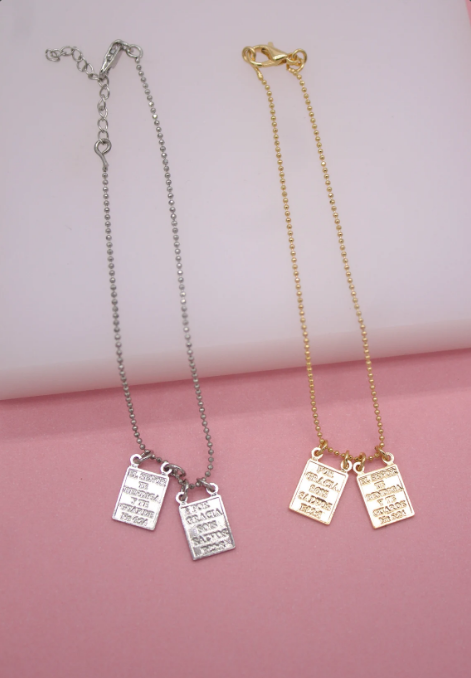 Two dainty ball chain bracelets, one silver and one gold, display rectangular engraved pendant charms featuring Spanish inscription details against a soft pink background.