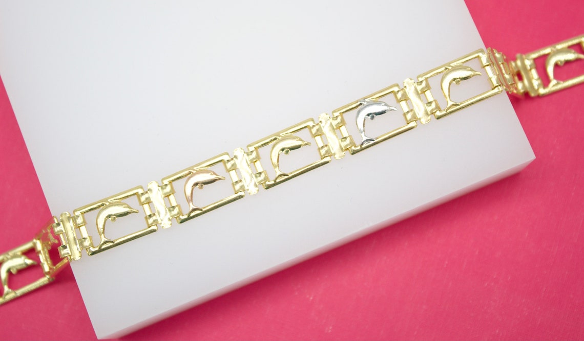 A tri-color gold dolphin bracelet rests diagonally across a white display block against a vibrant pink background.