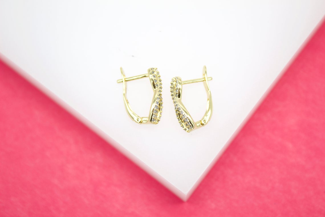 A pair of gold huggie hoop earrings embedded with sparkling diamonds rests on a white display surface against a bright pink background.