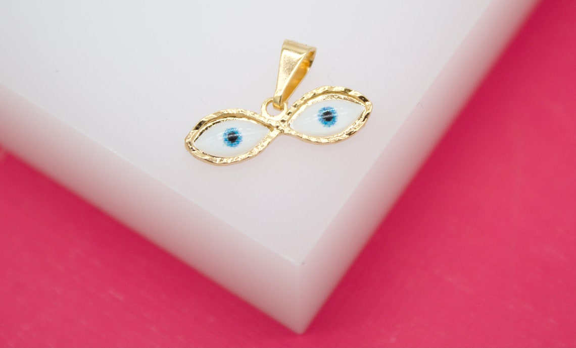 A gold evil eye pendant rests on a white display surface against a bright pink background.