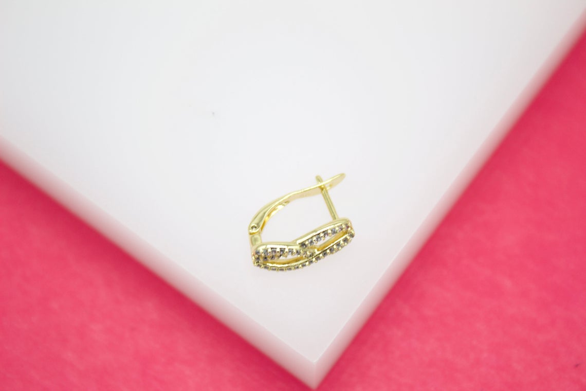 A gold-plated huggie hoop earring with embedded cubic zirconia stones rests on a white surface against a pink background.