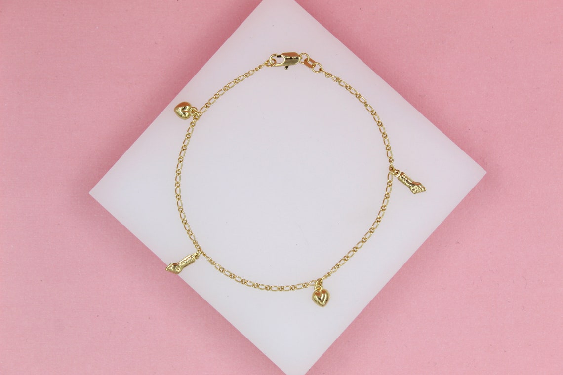 A delicate gold charm bracelet featuring alternating heart and high-heel shoe pendants rests on a white display against a pink background.