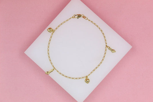 A delicate gold charm bracelet featuring alternating heart and high-heel shoe charms rests on a white display square against a soft pink background.