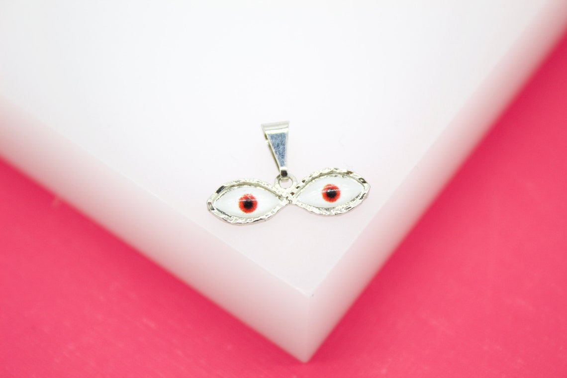 A silver double-eye pendant featuring white enamel and red pupils rests on a white display against a pink background.