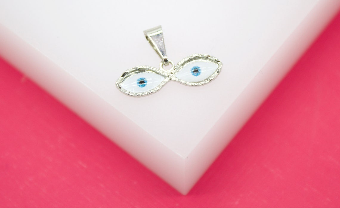 A silver evil eye pendant rests on a white display surface against a vibrant pink background.